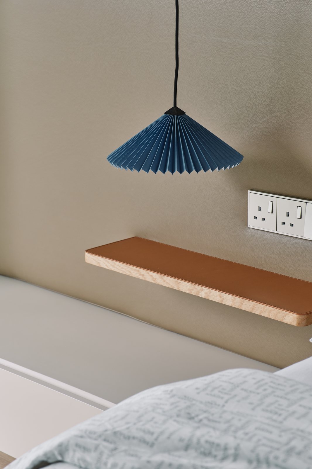 A bedside shelf in oak and leather, a pleated blue pendant above